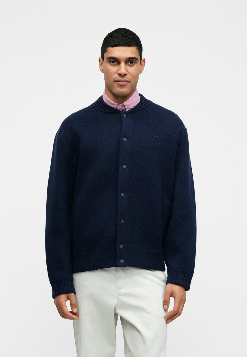 Tumesinine kudumärgiga kardigan ribatud krae, nupplukuga ja küljetaskutega. Kandmiseks helesinise särgi peal, millel on diskreetne logoelement.