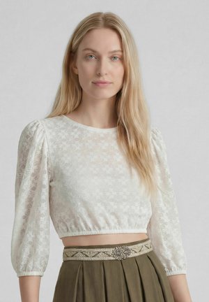 Blond vrouwtje met een witte gebloemde kanten cropped blouse met pofmouwen en een olijfgroene hooggetailleerde rok met decoratieve riem.