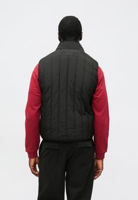 Gilet trapuntato nero con colletto alto, caratterizzato da cuciture verticali. Abbinato a una maglia rossa a maniche lunghe. Design semplice, senza elementi metallici visibili.