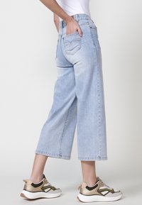 Los jeans de pierna ancha en un tono azul claro cuentan con una cintura alta, bolsillos traseros y un dobladillo deshilachado. Se combinan con zapatillas con detalles metálicos.