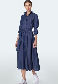 Dżinsowa sukienka koszulowa z długimi rękawami, kołnierzykiem, dwiema kieszeniami na piersiach, paskiem w talii oraz zwiewnej długości midi, zestawiona z białymi trampkami.