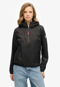 Veste légère noire avec capuche, fermeture éclair, deux poches latérales et poignets élastiques. Elle arbore un logo "Superdry" en rose sur la poitrine.