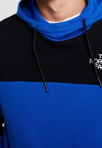 Modrá a černá mikina s kapucí, šňůrkou a měkkou texturou. Obsahuje design s barevnými bloky a logem na přední straně.