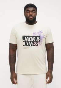 Cream-colored t-shirt with short sleeves, featuring a graphic design in black and purple, and "JACK & JONES" text prominently displayed.