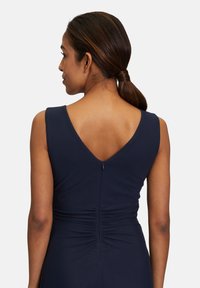 Navy mekko, jossa on V-mallinen selkäosa, istuva bodice ja rypytetty yksityiskohta vyötäröllä. Tasainen kangas, jossa on piilotettu vetoketjukiinnitys takana.