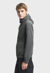 Grå hættejakke lavet af glat, let materiale. Har elastiske manchetter, en lynlåslomme og et subtilt logo på ærmet.