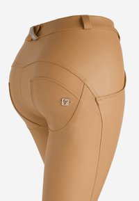 Leggings aderenti color tan con una texture liscia, cucitura curva sul retro e piccolo logo decorativo accanto alla tasca sinistra.