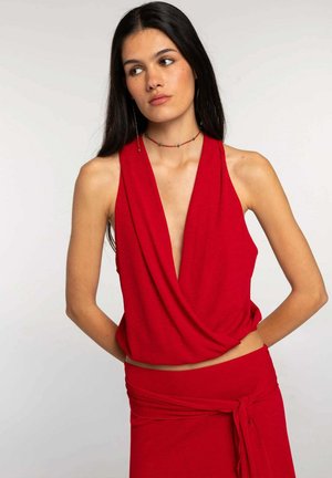 Joven mujer con cabello largo y oscuro que lleva un top rojo sin mangas con un pronunciado escote en V y una falda roja a juego atada a la cintura, mirando de lado.