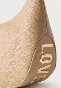 Γωνία τσάντας από μπεζ δερματίνη με ανάγλυφα χρυσά γράμματα που σχηματίζουν τη λέξη «LOVE» σε λευκό φόντο.