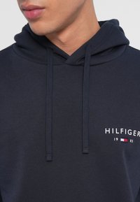 Námornícka mikina s kapucňou a sťahovacími šnúrkami, s logom "HILFIGER" a rokom "1985" v bielej farbe. Mäkká tkanina s textúrovaným povrchom.