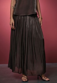 Faltenrock in Midi-Länge in Dunkelbraun, aus leichtem, glänzendem Stoff. Kombiniert mit einem passenden Oberteil und schwarzen Riemchensandalen.