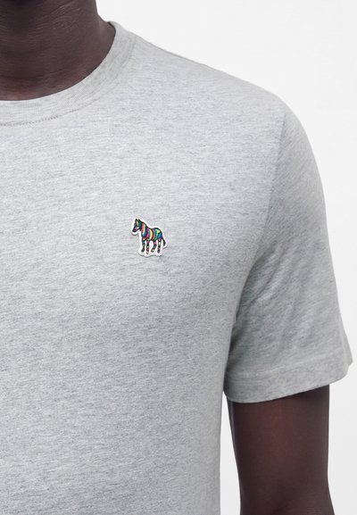 Personne portant une chemise à manches courtes gris clair avec un petit patch zèbre rayé multicolore sur la poitrine gauche.