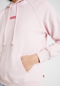 Sudadera rosa claro con capucha y cordones. Presenta un bolsillo frontal y un pequeño logo rojo de Levi's en el pecho. Tejido suave, puños acanalados.
