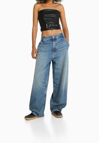 Top corto sin tirantes de cuero negro con un cierre en la parte delantera, combinado con unos vaqueros de denim azul claro de corte amplio y zapatillas negras con suelas de goma.