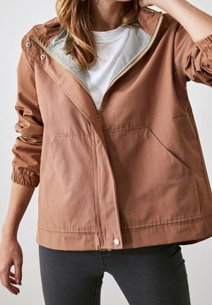 Vrouw draagt een lichtbruine jas met ritssluiting en elastische manchetten over een wit shirt en donkere broek, poseert met één arm omhoog bij het hoofd.