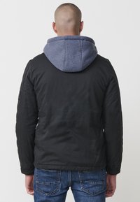 Melns uzvalks ar pelēku kapuci. Izceļas ar teksturētu audumu, vienkāršu dizainu un redzamām šuvēm. Apvienots ar zilām džinsu biksēm, redzams aizmugures skats.