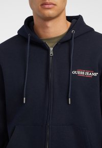 Navy Zip-Up Hoodie aus Baumwollmischung, mit Kapuze mit Kordelzug, Fronttasche und gesticktem Logo in Rot und Weiß auf der Brust.