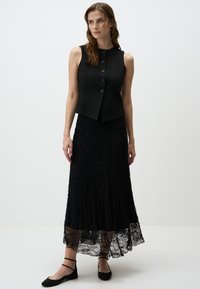 Top sans manches noir avec des boutons sur le devant associé à une longue jupe en dentelle noire. Présente un tissu texturé et une silhouette fluide. Chaussures plates noires.