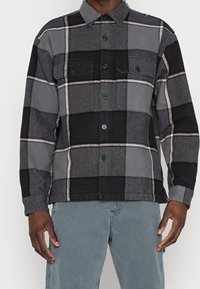 Musta ja harmaa ruudullinen flanellipaita, jossa on kauluksellinen pääntie, napitettava etuosa ja kaksi rintataskua. Pehmeä rakenne ja kontrastiset kuviot.