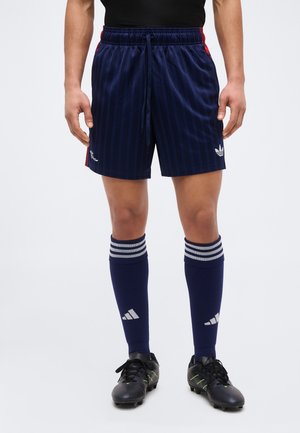 Άνδρας που φοράει σκούρο μπλε αθλητικό σορτς με το λογότυπο της Adidas, αντίστοιχες κάλτσες μέχρι το γόνατο με ρίγες και μαύρα ποδοσφαιρικά παπούτσια με τάπες.