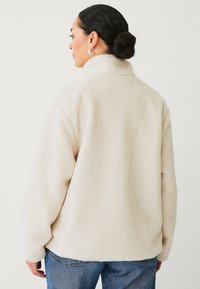 Beige fleecejakke med høj krave, sænkede skuldre og en blød tekstur. Båret med blå jeans, som viser en afslappet pasform.