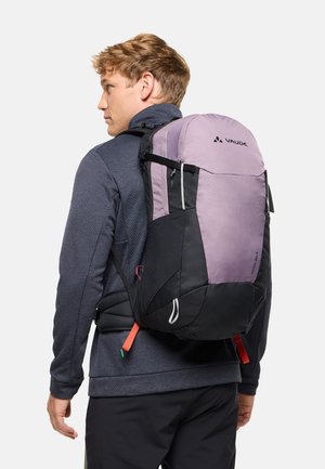 Jeune homme portant un sac à dos Vaude violet et noir avec des bretelles réglables, vêtu d'une veste texturée foncée, vu de profil sur un fond blanc.