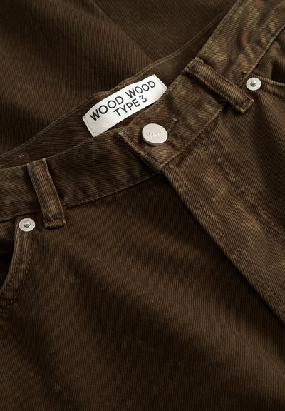 Pantaloni maro din catifea de tip șnur, cu o etichetă pe talie pe care scrie „WOOD WOOD TYPE 3”, dotată cu accesorii metalice argintii și o suprafață texturată a țesăturii.