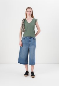 Oliven grønn ermeløs topp med blondedetaljer, bredbukser i blå denim og svarte sneakers med brune såler. Bærer en mønstrete veske.