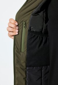 Mano sosteniendo una chaqueta verde abierta que muestra un bolsillo con cremallera y un bolsillo de malla negra en el forro acolchado.