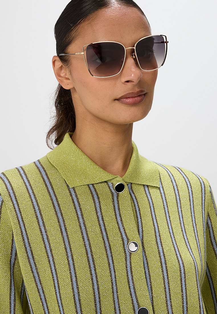 Camisa a rayas verdes con cuello, que presenta líneas verticales negras y azules, y grandes gafas de sol con lentes marrones degradados.