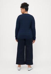 Ναυτικό πλεκτό πουλόβερ με υφή, συνδυασμένο με σκούρες φαρδιές τζιν και ασημένιες μπαλαρίνες. Άποψη από πίσω, δείχνοντας το ριμπέ χείλος και τα μανίκια.