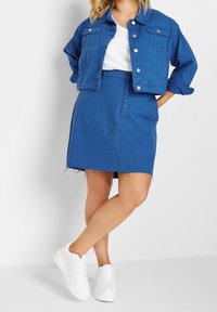 Modrá džínsová bunda s krátkým střihem, dvěma předními kapsami a vyhrnutými rukávy, v kombinaci s odpovídající modrou džínsovou sukní a bílými teniskami.