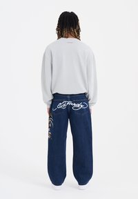 Lys grå sweatshirt med lange ærmer, parret med løse blå jeans med hvidt skrifttype-logo og farverig sidebroderi.