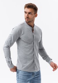 Camisa gris de manga larga con botones y un cuello mao, decorada con ribetes rojo, blanco y azul. Confeccionada en una tela suave y con un diseño ajustado.