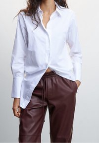 White button-up shirt with a pointed collar and long sleeves, worn over burgundy leather jogger pants with a drawstring waistband.