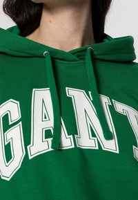 Gros plan d'un sweat à capuche vert avec des lettres blanches en bloc formant "GANT" et des cordons verts suspendus à la capuche.
