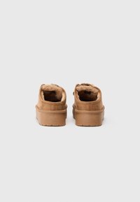 O pereche de pantofi slip-on din suede bej, cu talpă groasă și căptușeală pufoasă din blană, prezentată din spate pe un fundal simplu deschis.