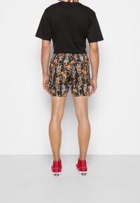 Hombre vistiendo una camiseta negra, pantalones cortos con un patrón floral en naranja y negro, y zapatillas rojas de caña alta, de pie frente a una pared blanca.