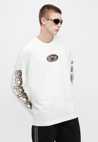 Fehér hosszú ujjú póló lila és fekete nyomott logóval a mellkasán és "adidas" felirattal az ujjakon; karcsúsított fekete farmernadrággal és napszemüveggel viselve.