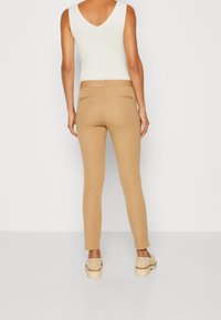 Pantalon beige ajusté avec une texture lisse. Dispose de deux poches arrière et d'une taille mi-haute. Assorti à un haut sans manches crème.