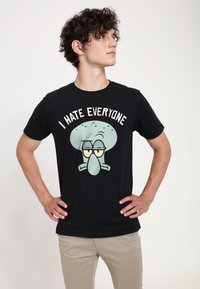 Sort bomulds t-shirt med en tegneseriefigur med et lyseblåt hoved og udtryk, samt teksten "JEG HADER ALLE" ovenfor.