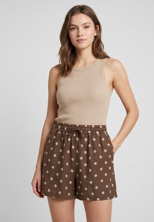 Junge Frau trägt ein beiges, geripptes ärmelloses Top und braune, hoch taillierte Shorts mit beigen Punkten, steht mit einer Hand in der Tasche.