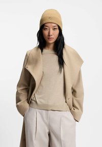 Beige ribbad neulakk, jossa on logo, tanvärinen kerrospaita ja beige takki avoimella kauluksella, yhdistettynä vaaleisiin, laskostettuihin housuihin.