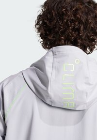 Lichtgrijze hooded jacket met reflecterende groene "CLIMA" tekst, textuurpaneel op de schouder en contrasterende stikdetails.