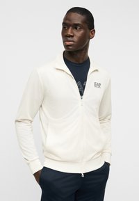 Ζακέτα με φερμουάρ σε κρεμ ύφασμα, με υφή στην επιφάνεια, γιακά, πλαϊνές τσέπες και λογότυπο EA7 στο αριστερό στήθος. Φορέθηκε πάνω από πουκάμισο σκούρου μπλε χρώματος.