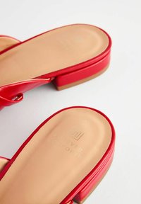 Tongs synthétiques rouges avec une semelle plate, présentant une texture lisse et un logo en relief sur la semelle intérieure. Design simple avec des accents minimes.