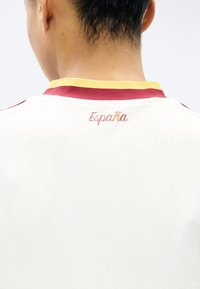 Πλάτη ατόμου με λευκή αθλητική φανέλα με κόκκινο και κίτρινο γιακά, με την επιγραφή "España" σε κόκκινο και κίτρινο στο πάνω μέρος της πλάτης.