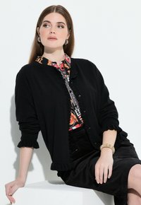 Černý cardigan s volánovými manžetami, zapínáním na knoflíky, přeložený přes barevnou vzorovanou blůzu. Modelka zkombinovala s černou sukní a strukturovaným náramkem.
