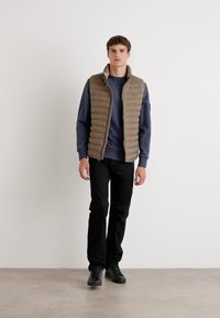 Gevulde bruine vest over een marineblauw sweatshirt, zwarte broek en zwarte schoenen. Kenmerkt zich door een hoge kraag en verticale stiksels op het vest.