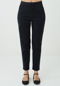 Pantalons noirs taille haute associés à des ballerines noires, dotés d'une simple fermeture à bouton et à zip, présentés sur un fond neutre.
